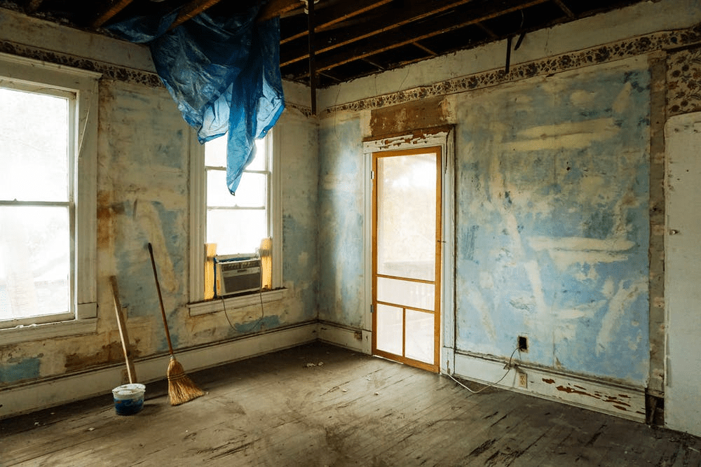 The interior of a distressed home. The walls have been stripped bare, there is a tarp handing from the ceiling rafters, and it is very dirty.